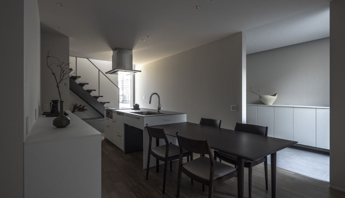 ダイニングテーブルとアイランドキッチンを直線的に配置し、奥に階段が見えるミニマルなLDKの様子